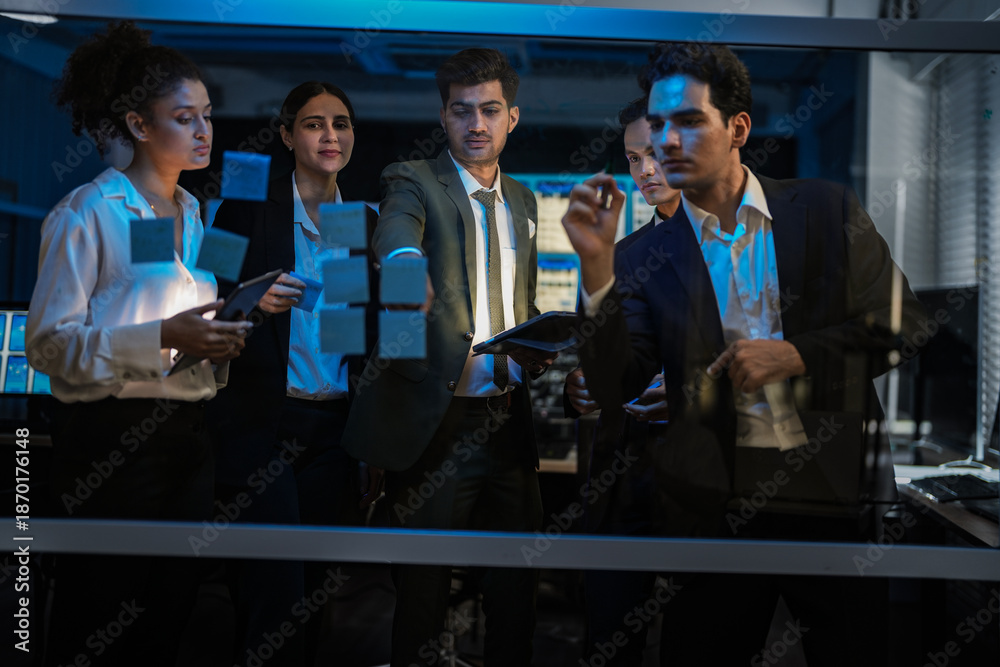 Obraz premium Agile tech startup team conducting scrum meeting on glass wall. Developers and managers mapping software workflow with sticky notes in modern blue-lit office at night.