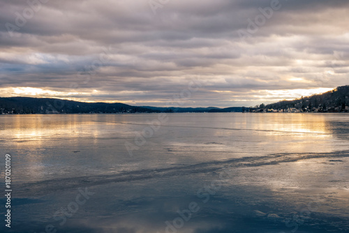 Calm winter view across Greenwood Lake, NY, with smooth ice and golden reflections beneath heavy clouds, stretching toward distant hills and lakeside homes in the quiet cold of winter.