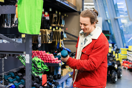 Fotografie Smiling man in red jacket holding blue dumbbell while shopping for sports equipm