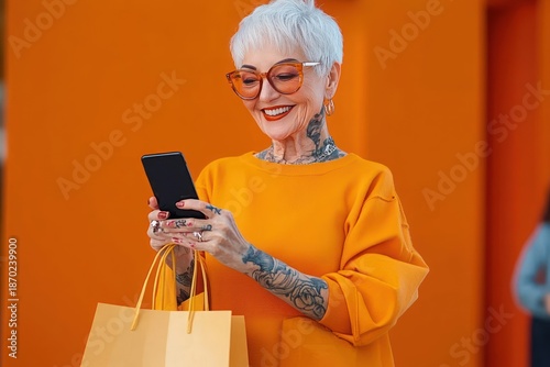 Wallpaper Mural mature white-haired woman in bright orange sweater with visible tattoos and hoop earrings holding a yellow shopping bag and using a smartphone against vivid orange background, focused Torontodigital.ca
