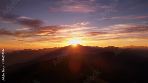 A breathtaking time-lapse sequence captures the majestic beauty of a sunrise or sunset over a sprawling mountain range. Golden, warm light dramatically bursts from behind a prominent peak, casting vib