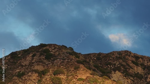 Dramatic view of sunlit coastal hills beneath heavy blue storm clouds, highlighting rugged terrain, sparse vegetation, and shifting light before rain.