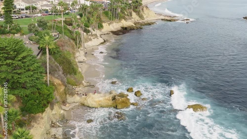 Aerial view of a calm coastal bay with palm trees, rocky shoreline, and hillside homes stretching along the coast. Gentle waves roll onto small coves, creating a relaxed seaside atmosphere.