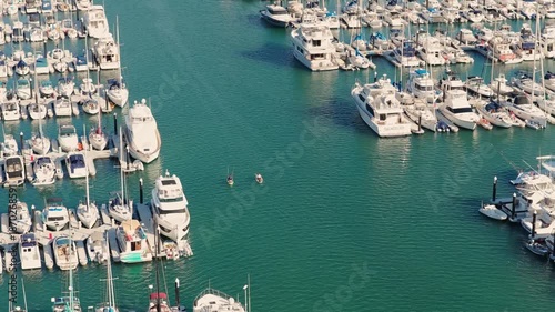 A calm aerial view over a marina: rows of white yachts docked on both sides, clear turquoise water in the center, and two kayakers moving quietly through the harbor.