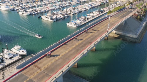 Aerial view of cyclists crossing a bridge over a marina filled with docked boats, with calm turquoise water and a small motorboat passing below on a sunny day.