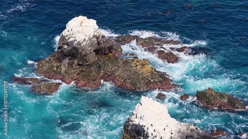 Aerial view of rugged sea rocks rising from deep blue water, with white waves crashing around the formations and turquoise currents swirling along the rocky shoreline.