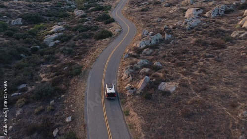 Aerial view of a lone vehicle driving along a winding road through a dry, rocky landscape with rolling hills and distant mountains under soft evening light.