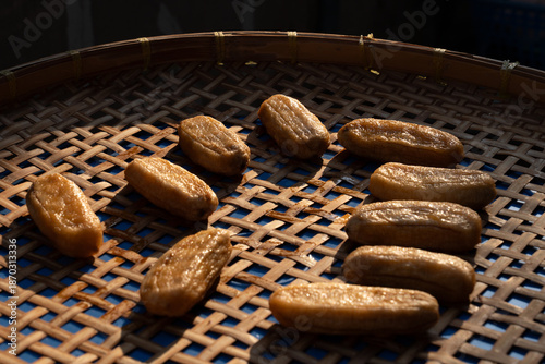 Low-angle perspective shot of peeled bananas drying on a bamboo sieve under strong sunlight. The image features high contrast with deep shadows in the background.