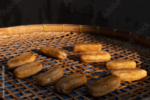 Perspective view of sun-dried bananas arranged on a woven bamboo tray, illuminated by sunlight against a dark background.