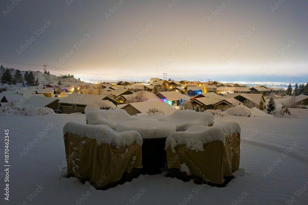 Fototapeta premium View from a backyard patio with covered furniture of Illuminated homes in a hilltop subdivision above the lights of the city of Spokane, Washington on a winter evening with snow on the ground.