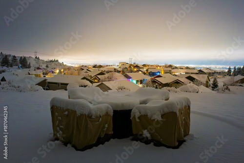 View from a backyard patio with covered furniture of Illuminated homes in a hilltop subdivision above the lights of the city of Spokane, Washington on a winter evening with snow on the ground.