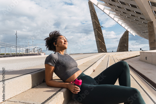 Woman in great shape taking a break on steps, enjoying music and staying hydrated after outdoor workout