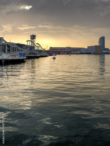 Sunset over Barcelona waterfront with boats, golden sky, and iconic modern architecture. Sunset at Barcelona Port Vell with Rambla de Mar bridge and W Hotel under a dramatic cloudy sky.
