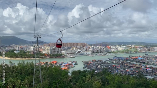 Red cable car cabin riding along ropeway above busy fishing harbor toward Nanwan Monkey Island, with coastal city skyline and mountains in background — Xincun, Hainan, China.
