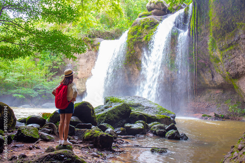 Valokuva Female tourist wearing a hat and carrying an adventure backpack stands in front of Heo Suwat Waterfall, Khao Yai National Park, Nakhon Ratchasima, Thailand