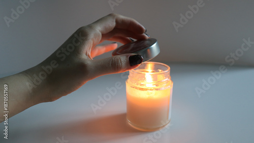 Womans hand covers a burning scented candle with lid, extinguishing the flame. The candle's light creates warm, cozy atmosphere. Idea for meditation, spa procedures, self care, setting romantic mood.