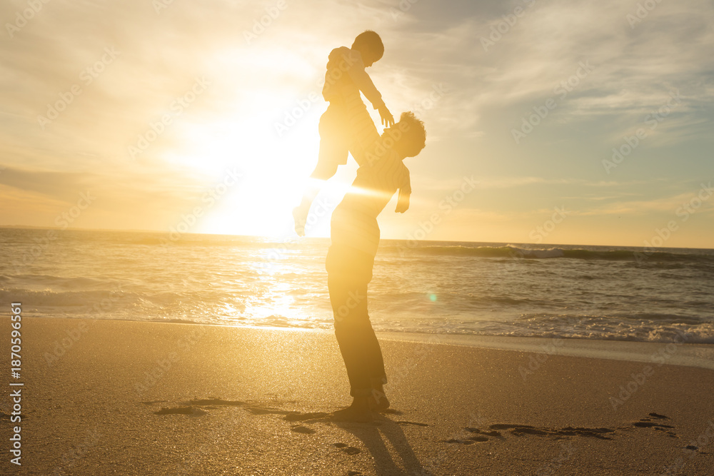 Fototapeta premium Side view of back lit father holding aloft son on shore at beach against sky during sunset