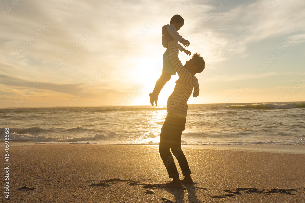 Fototapeta premium Side view of playful father holding son aloft on shore at beach against sky during sunset