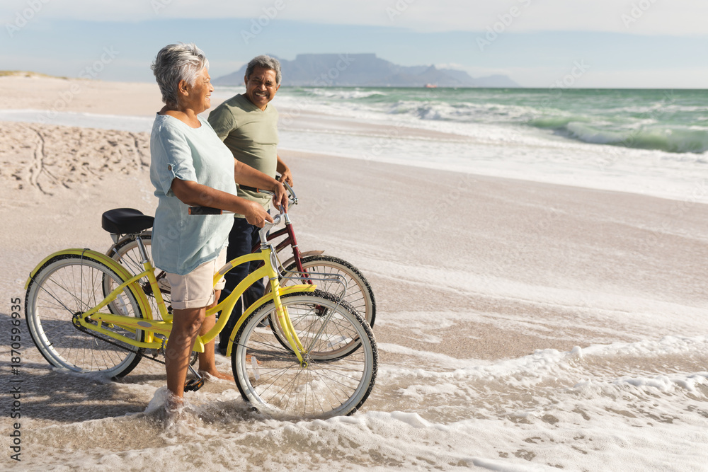 Fototapeta premium Full length of senior multiracial couple with bicycles standing on shore at beach during sunny day