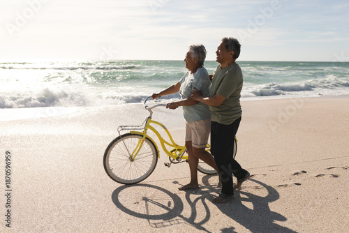 Side view of multiracial senior couple walking with bicycle on shore at beach during sunny day
