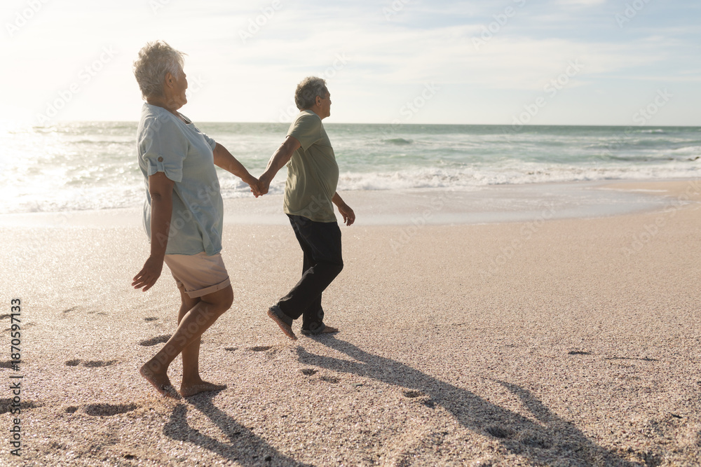 Fototapeta premium Full length side view of multiracial senior couple holding hands while walking on shore at beach