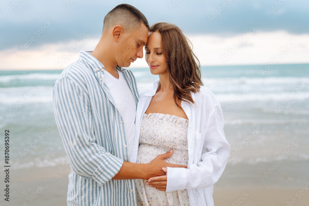 Obraz premium Happy expectant parents stand on the beach and enjoy the seascape. A man and his pregnant wife stroll along the seashore. Concepts of family, expectation, and love.