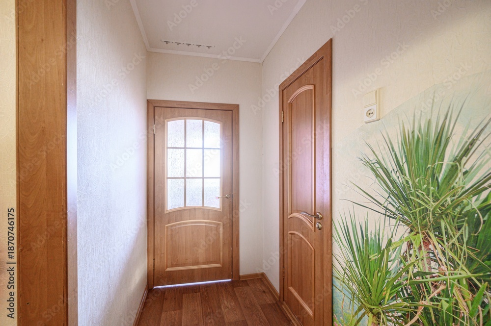 Fototapeta premium A narrow hallway with light wooden doors (one with frosted glass panels), wooden flooring, beige walls, and a large botanical mural on the right wall. Ceiling spotlights provide illumination