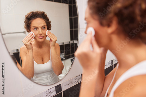Εκτύπωση καμβά A woman with curly hair is using a cotton pad to cleanse her face in front of a bathroom mirror