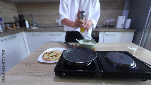 A person cooks pancakes on a portable electric stove. After flipping a golden-brown pancake onto a plate, they grease the pan and pour fresh batter to start the next one.