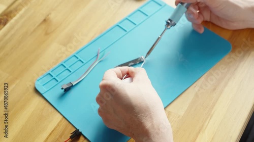 Top view close-up of hands soldering a small circuit board, one holding solder wire and the other a soldering iron, applying tin on contacts on a silicone mat