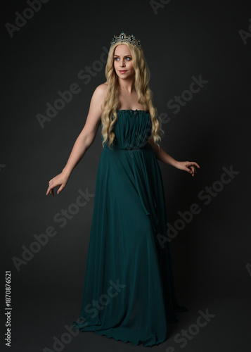 Full length portrait of blonde female model wearing royal green ballgown, fantasy crown. Standing, walking  pose, front view isolated figure on dark studio background.