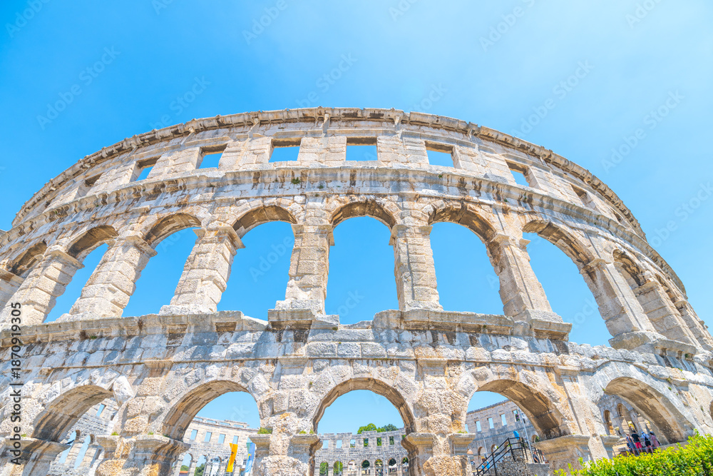 Naklejka premium This stunning Roman amphitheatre in Pula stands majestically under a clear blue sky, showcasing its impressive architecture and historical significance as a cultural landmark.