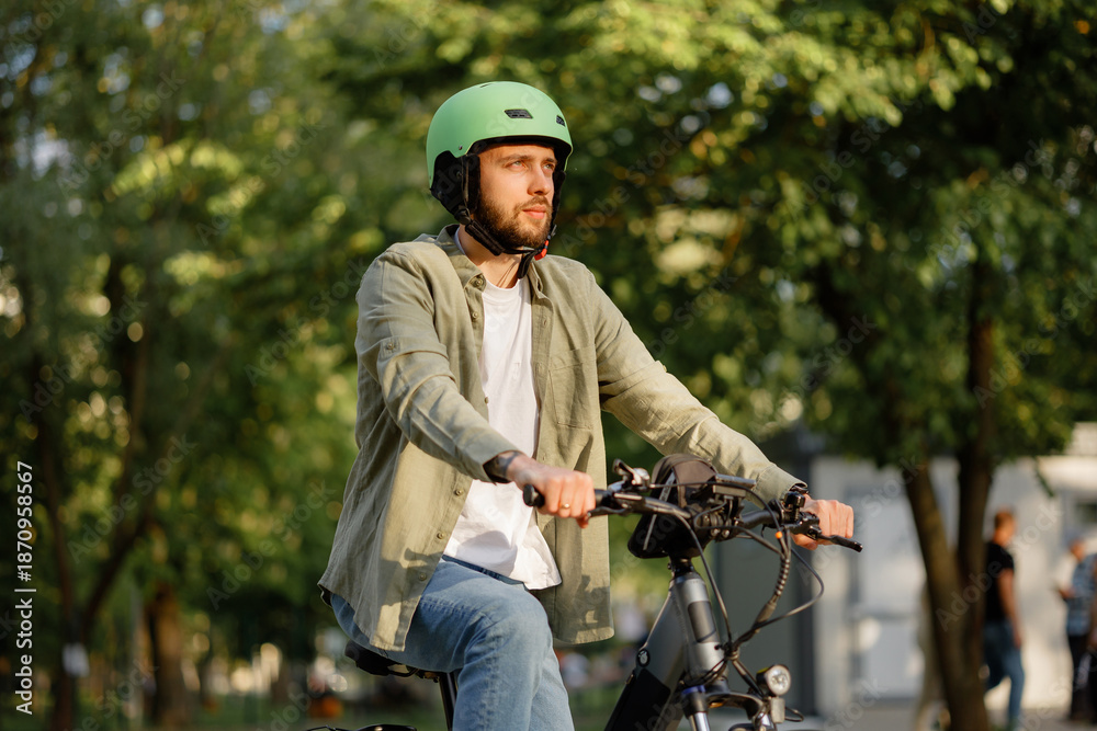 Fototapeta premium Man rides bicycle in city park during sunny day with bright green helmet and casual clothing while trees surround him