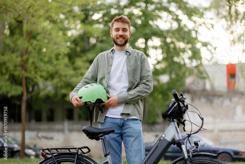 Young man holds a green bicycle helmet while standing next to his bike in a park during daytime