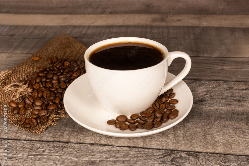 Fototapeta premium Black coffee in white ceramic cup with coffee beans on burlap sack and wooden background. 