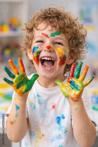 Joyful child with colorful paint smudges on face and hands laughs excitedly. Curly hair kid expresses happiness, playing with vibrant art supplies indoors.