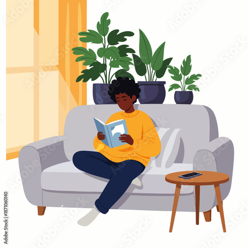 Young African American man relaxing at home on a comfortable sofa, engrossed in reading a book, surrounded by green houseplants, enjoying quiet leisure time indoors.