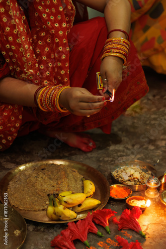 Indian woman in red sari wearing colorful stacked glass bangles performs puja with decorated thali lighting incense sticks with flowers banana diya and pooja offerings around