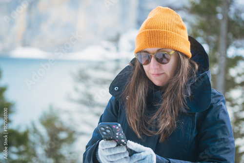 Young woman in winter clothing and sunglasses looking down at her smartphone screen in a snowy landscape