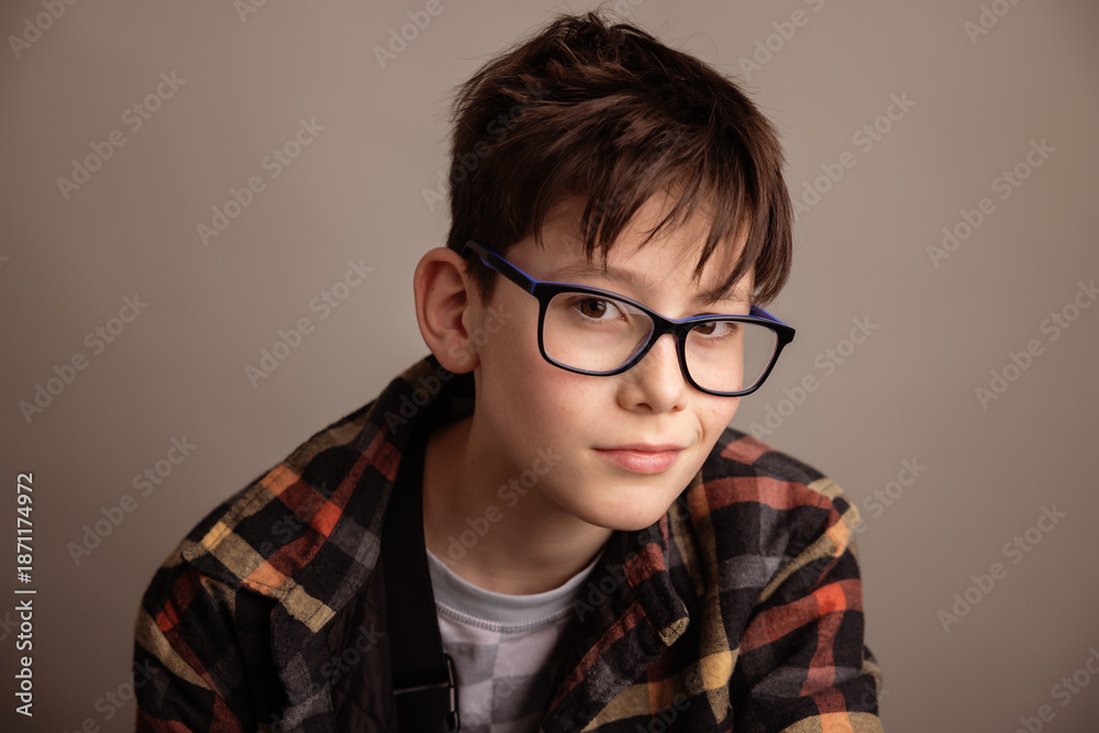 Fototapeta premium Thoughtful studio portrait of a young boy wearing eyeglasses and a casual plaid shirt, neutral beige background, soft natural light