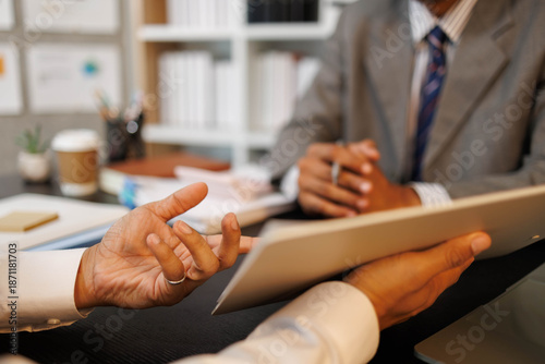 
Two men are sitting at a desk with a tablet in front of them. One of the men is holding the tablet and the other is looking at it. The scene suggests a meeting or discussion between the two men
