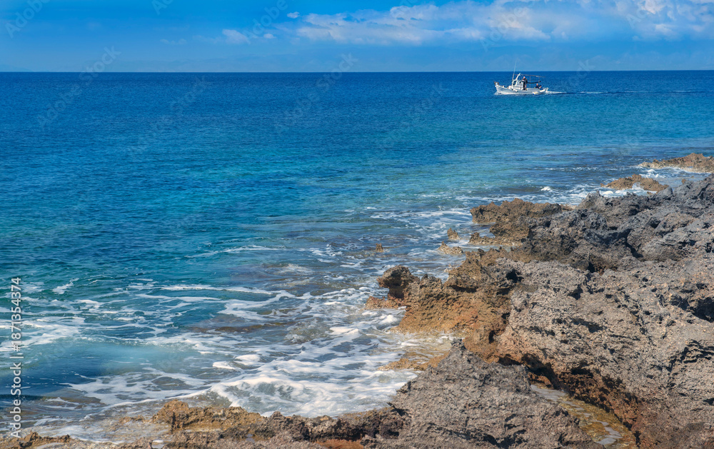 Fototapeta premium sea view from rocky coast with a beautiful blue water and a fishing boat in the distance in Peloponnese in Greece