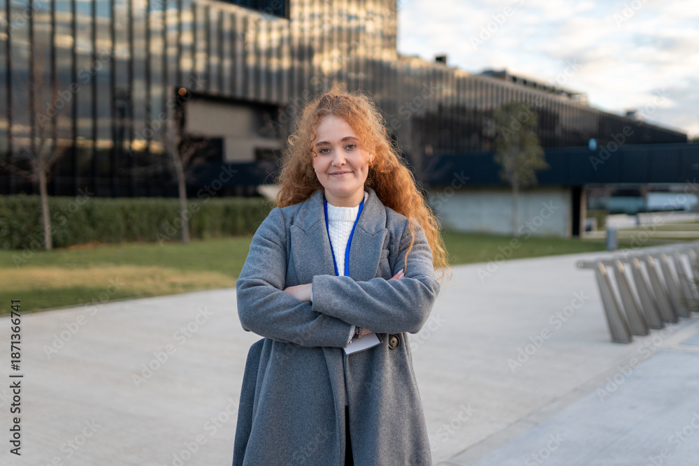 Fototapeta premium Red haired businesswoman standing with crossed arms in front of corporate building, showing confidence and professionalism