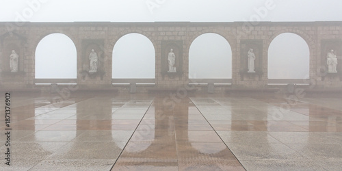 Square of Montserrat abbey on a rainy and foggy day, with a wall with arches and statues reflecting in the wet pavement stones. Catalonia, Spain 