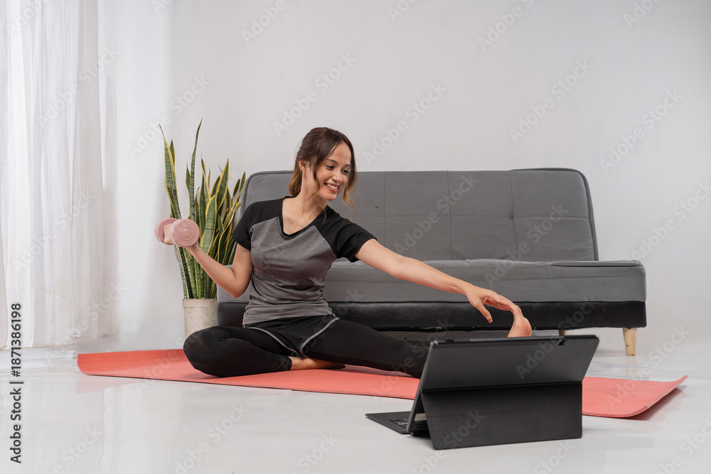 Fototapeta premium Asian trainer holds small dumbbell on mat, seated twist for tone and mobility with laptop for virtual coaching, compact home