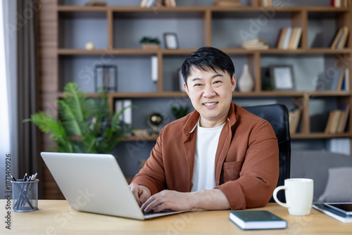 Φωτογραφία Smiling asian man in smart-casual attire types on a laptop at a wooden desk in a