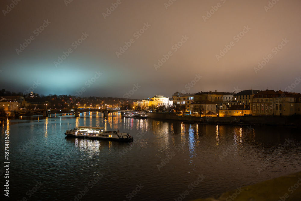 Fototapeta premium Prague city at night with illuminated buildings and a boat on the river Vltava during December. Cityscape of the historical capital of Bohemia in the Czech Republic.