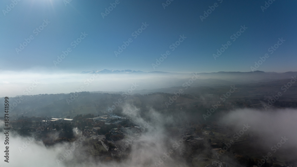 Obraz premium Aerial drone photograph revealing a stunning landscape of rolling hills and cultivated fields in the Marche region of Italy, partially covered by soft morning fog.