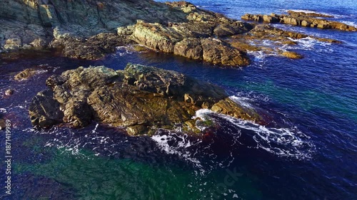 Coastal rocks rise majestically from crystal-clear waters, with waves dancing around them under the warm embrace of midday sun, creating a peaceful coastal scene.