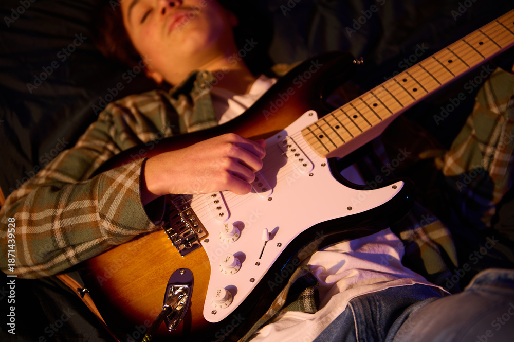 Obraz premium Relaxed musician lies back and strums an electric guitar in a warm, intimate setting. Plaid shirt and soft lighting convey a casual, cozy vibe for music lifestyle themes.